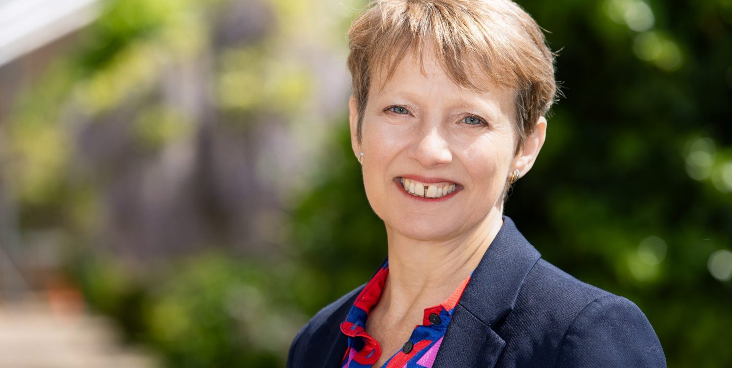 PCC Smiling, wearing a navy jacket and pink patterned shirt. It is sunny and the background is blurred foliage.