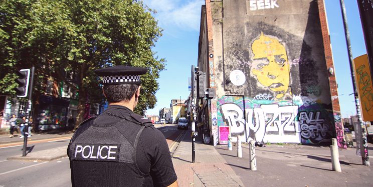 A police officer looks down a city street. A green tree, road and graffitied building are visible.